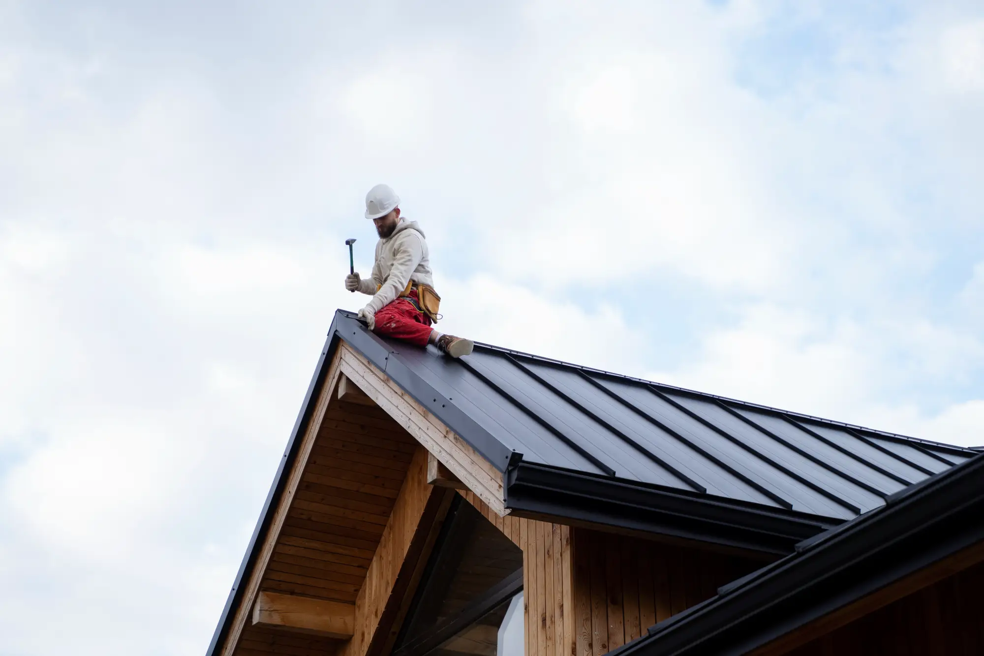 A construction worker in a white hard hat and red pants is sitting on the black metal roof of a modern wooden house, holding a hammer