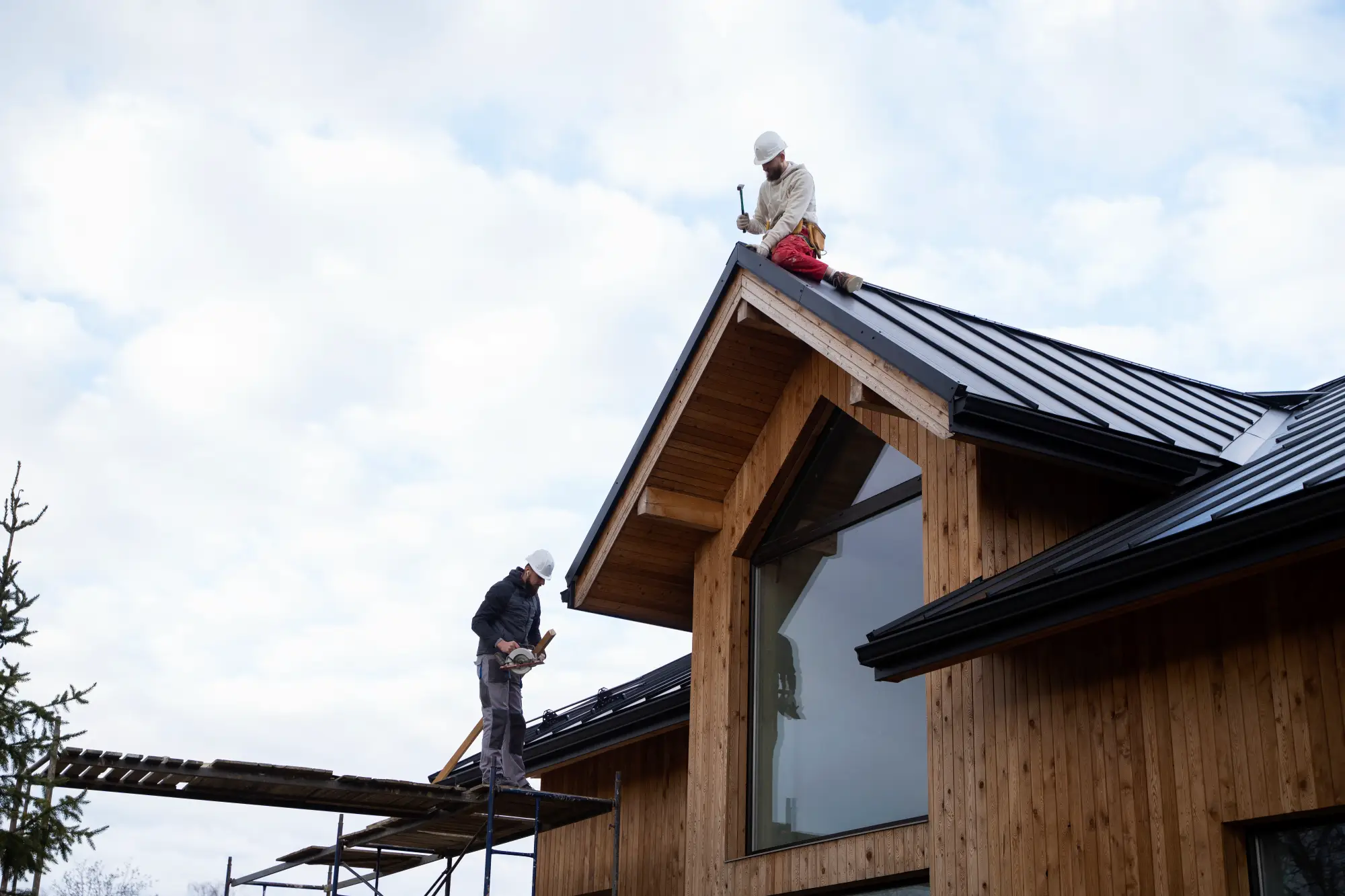 Two construction workers in hard hats are on the roof of a modern wooden house with a black metal roof