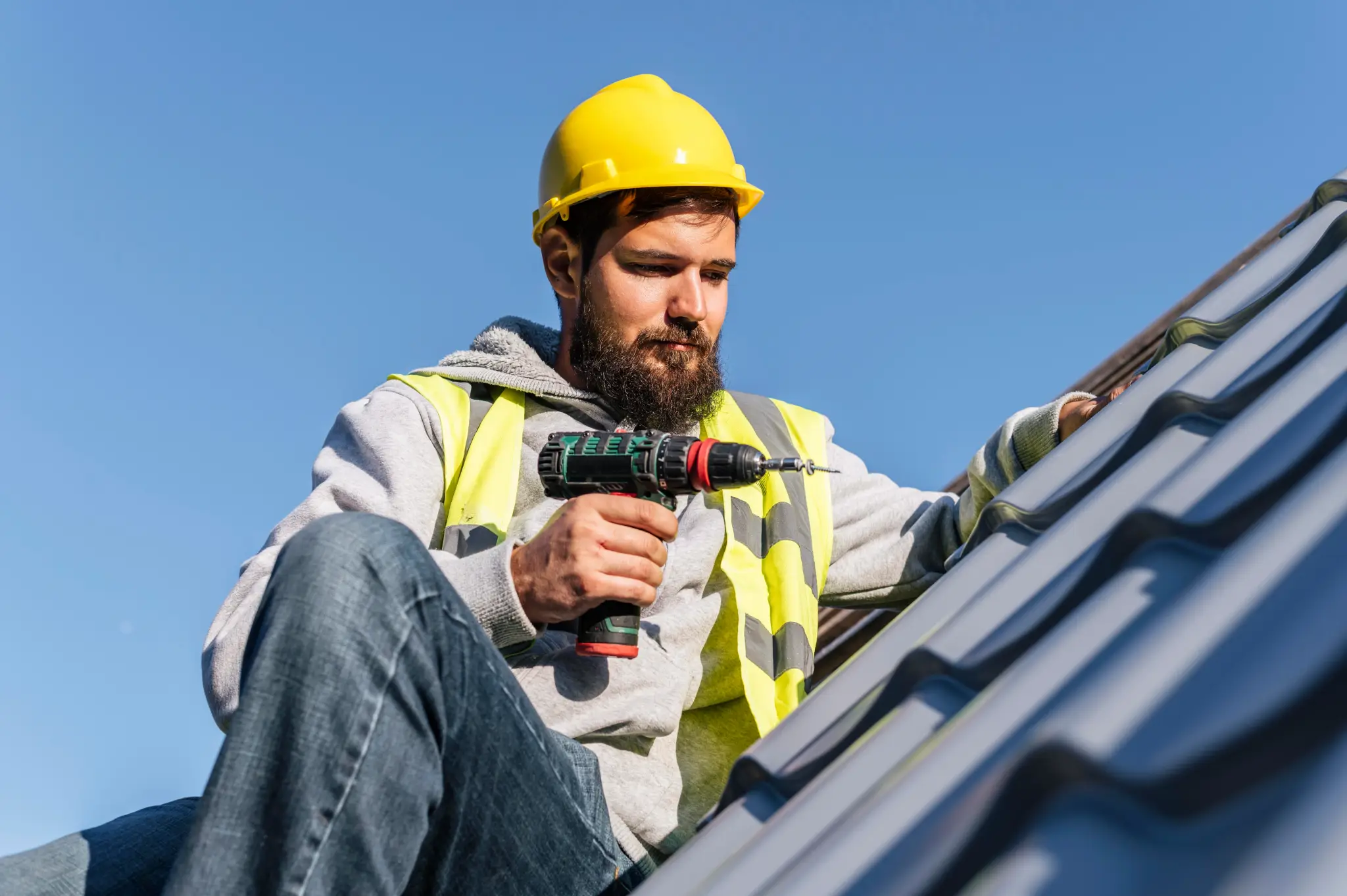A man with a beard and a yellow hard hat is on a roof, using a power drill on a gray metal roof