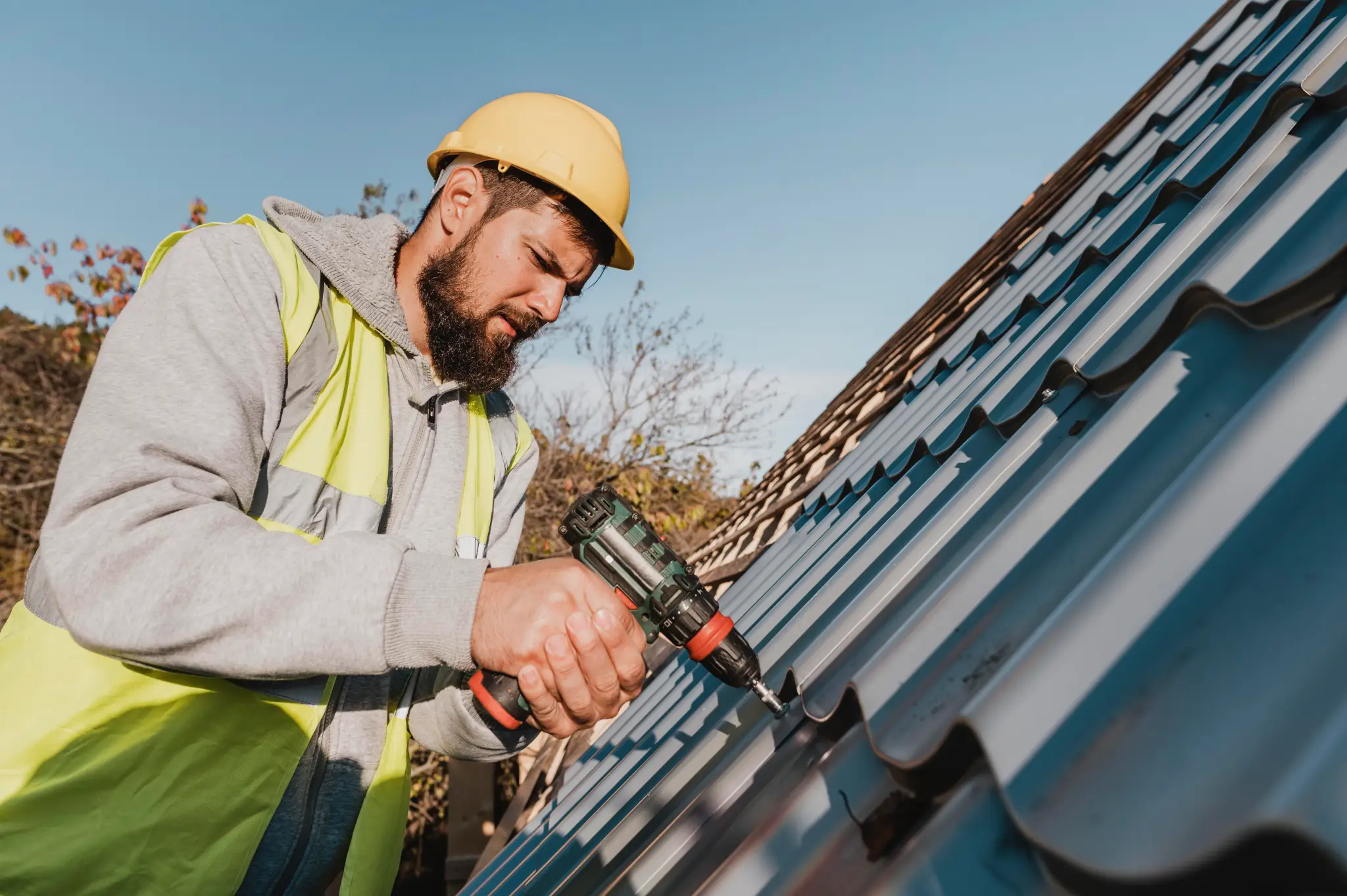 A man with a beard and a yellow hard hat is using a power drill on a gray metal roof