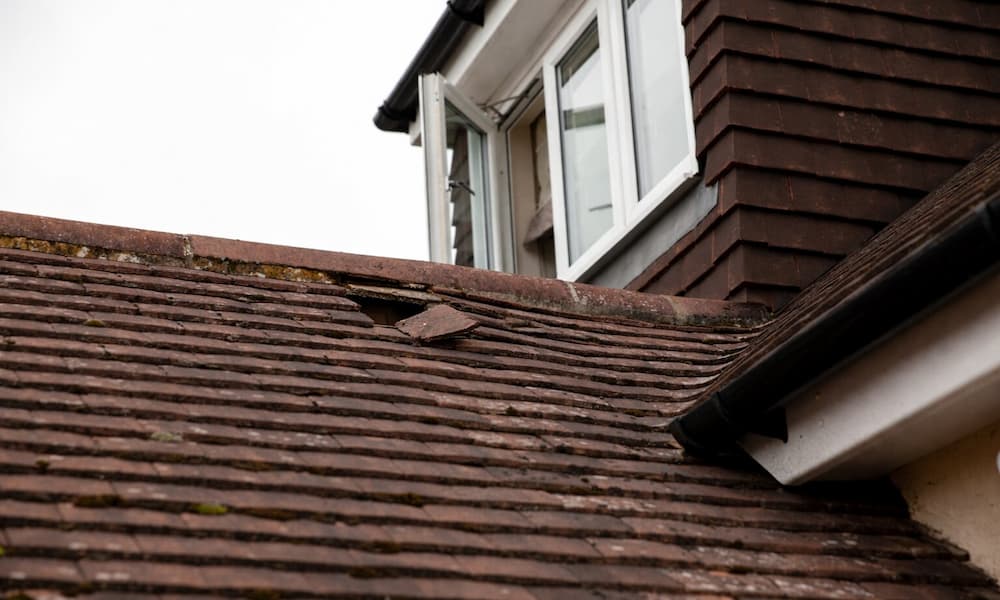 “Broken roof shingles with missing tiles exposing underlayment near window